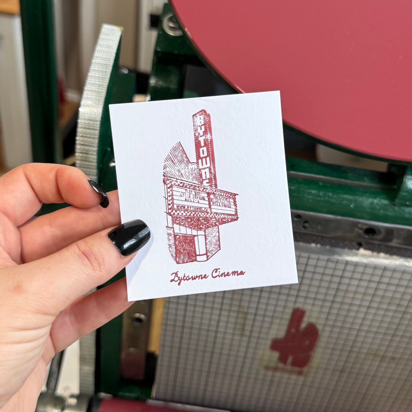 Hand holding a card with a red and white graphic design of a building labeled 'Bytowne Cinema' in front of a printing press.