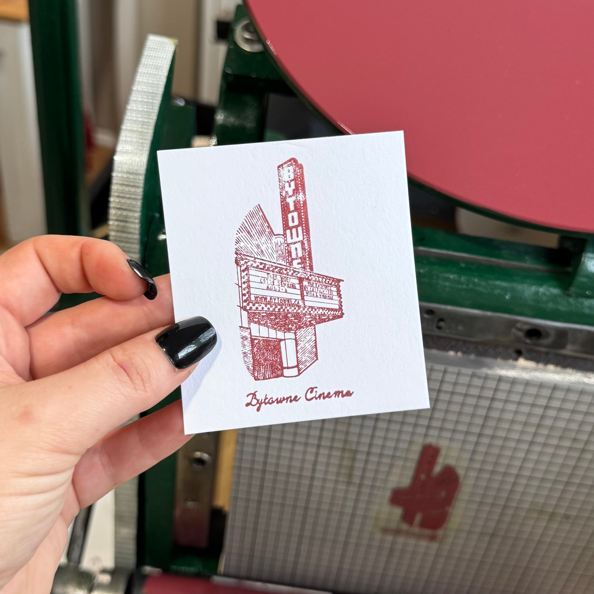 Hand holding a card with a red and white graphic design of a building labeled 'Bytowne Cinema' in front of a printing press.