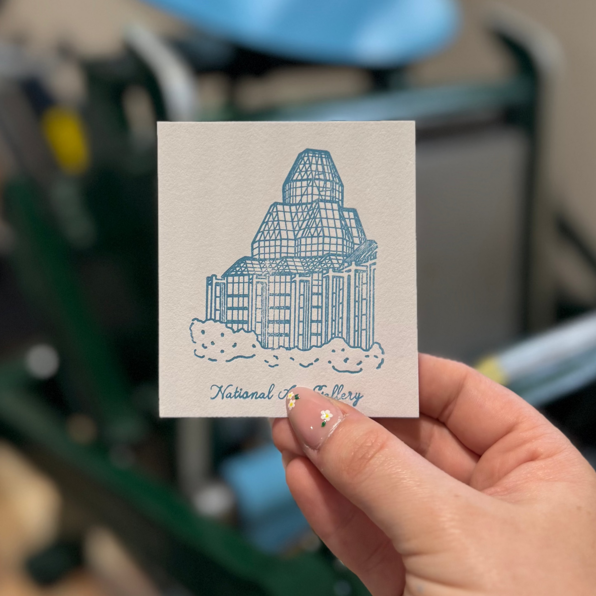 Hand holding a small card with a building design in front of a letterpress machine