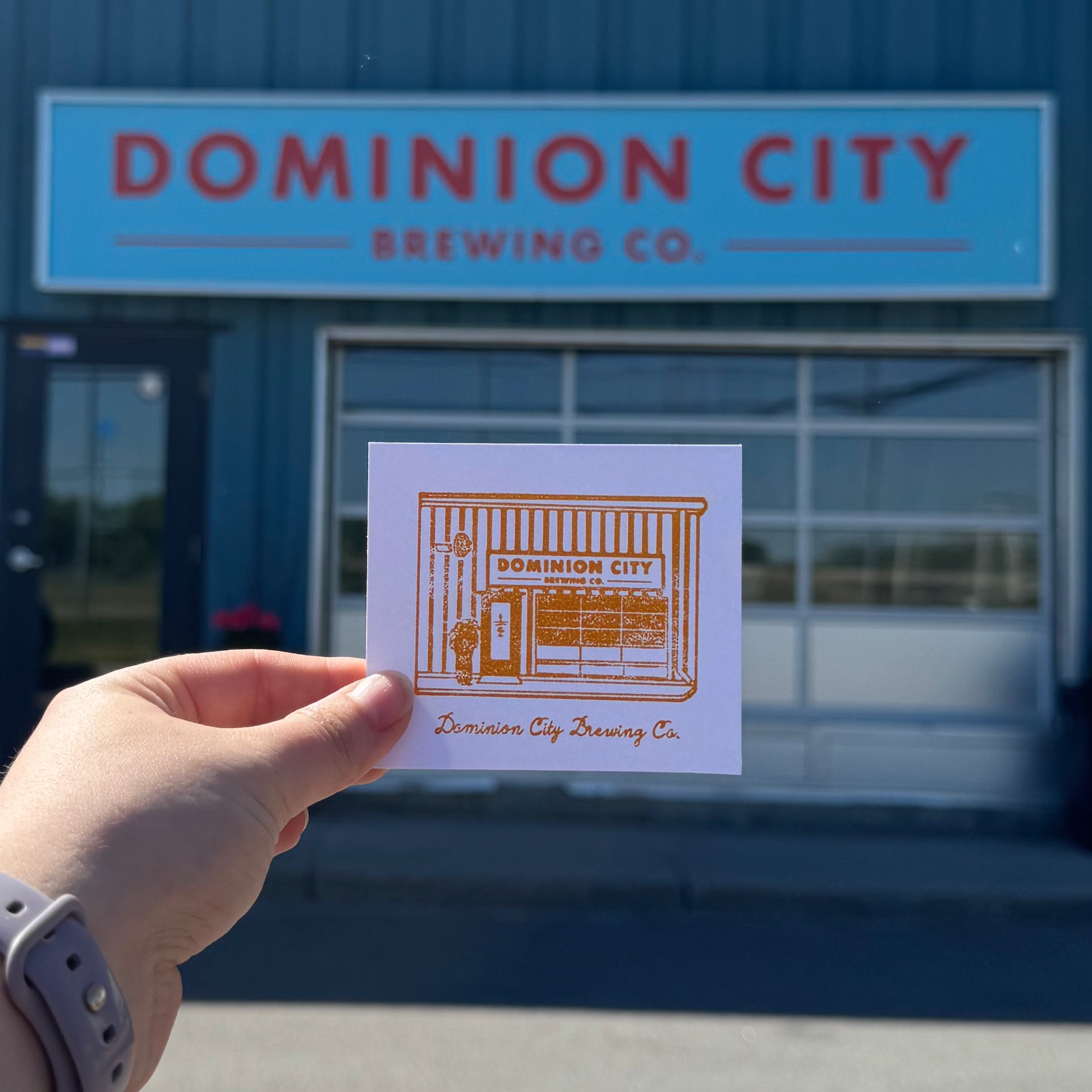 Hand holding a Dominion City Brewing Co. business card in front of the brewery building.