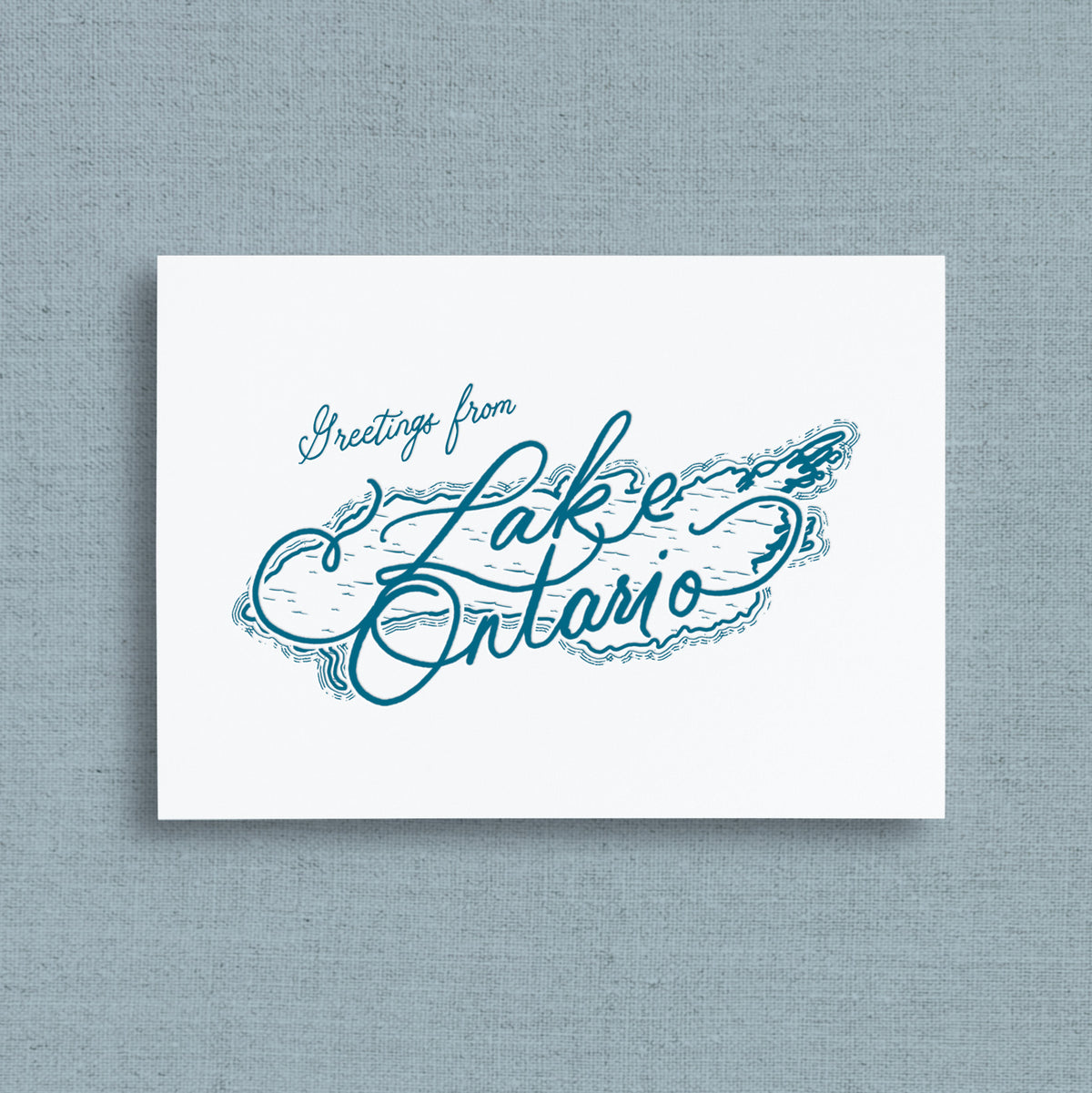 A white greeting card with blue script reading 'Greetings from Lake Ontario,' shown against a textured light blue background.